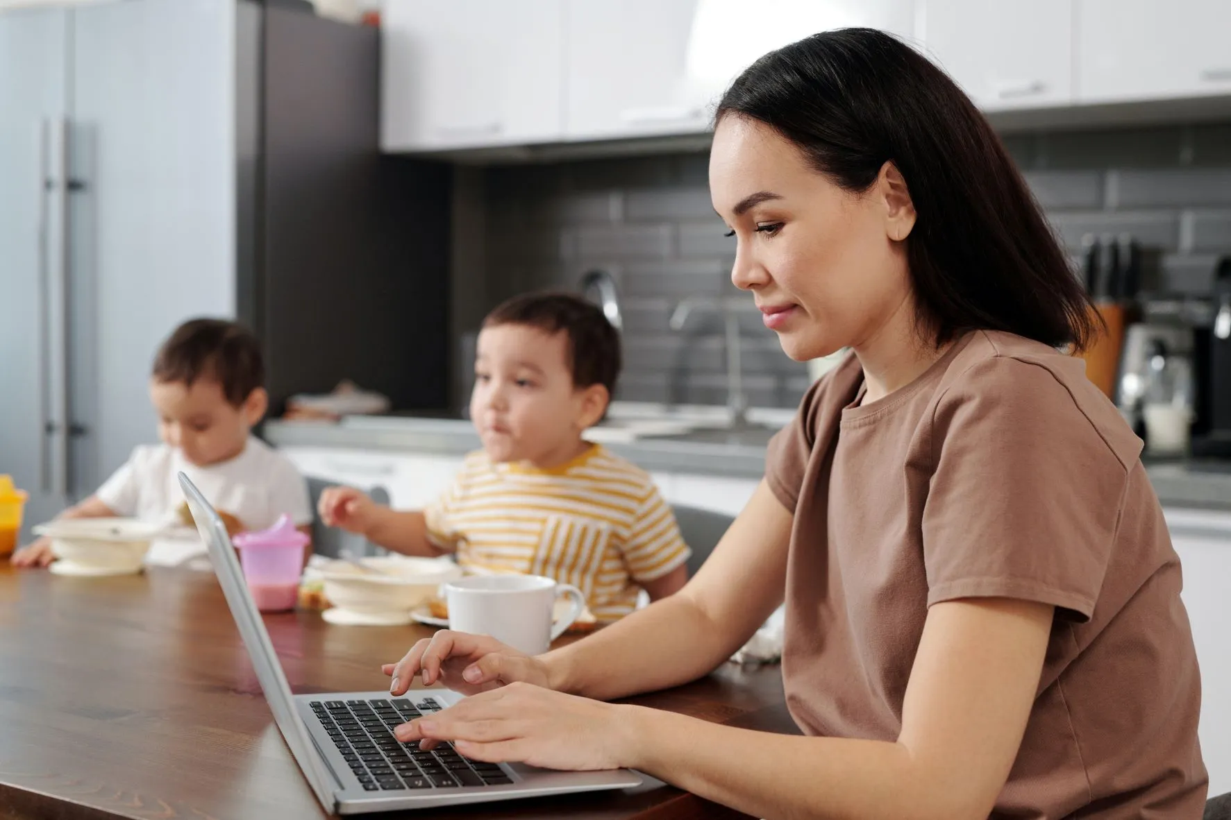 Workplace flexibility is depicted in an image showing a mother working on her laptop, in her family room/kitchen, with her two children in the background having food.