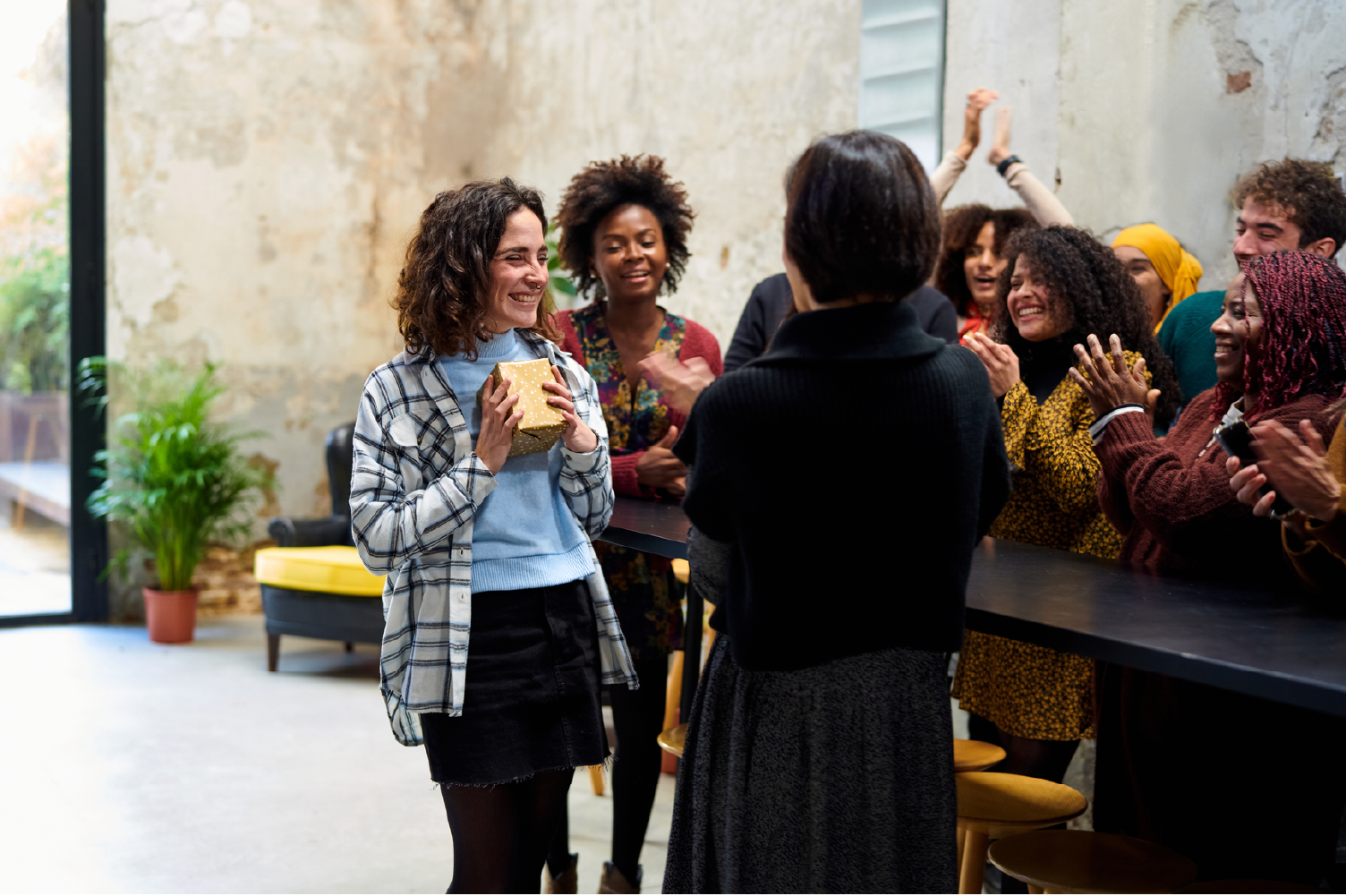  Languages of appreciation in the workplace is demonstrated with a woman in an office holds a gold-wrapped gift. Her colleagues are huddled around her, congratulating her on her work