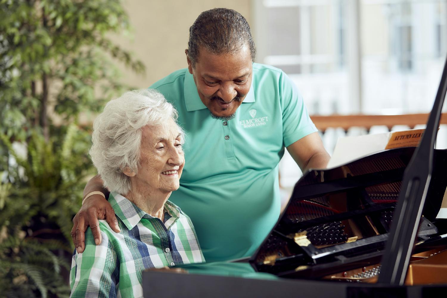 A caregiver socializes with a resident as she plays the piano.