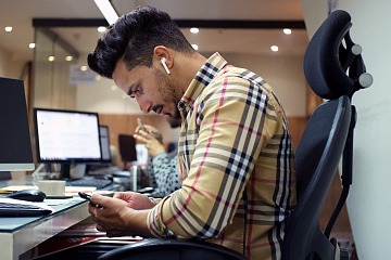 Employee wearing headphones looks at his phone while sitting at a desk